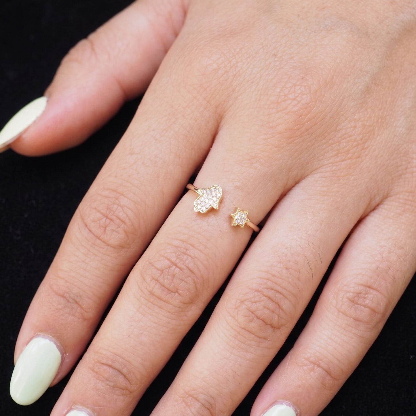 A close-up of a hand wearing two delicate gold rings against a black background. The larger ring features a textured, flower-like cluster of small clear stones, while the smaller ring is a simple gold band with a tiny clear stone.