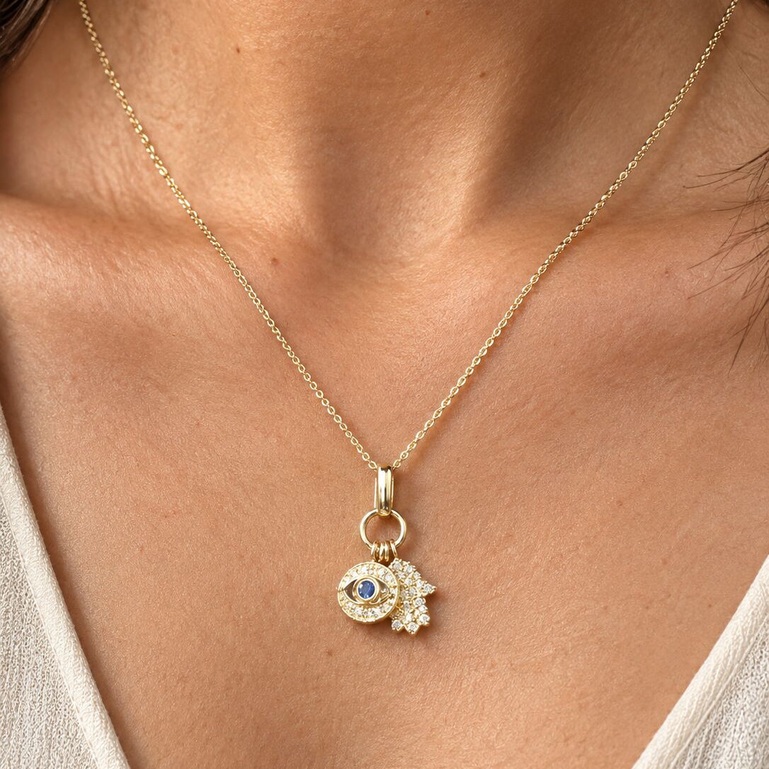 A close-up of a model wearing a delicate gold chain necklace with a double charm pendant. The pendant features a small gold evil eye with a blue gemstone and a gold hand-shaped charm paved with clear stones.