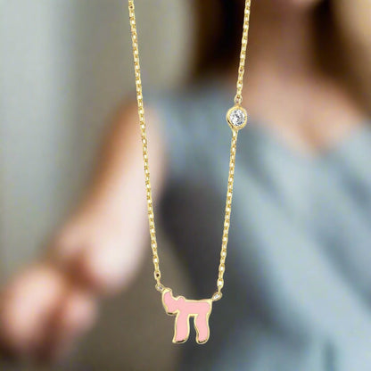 A Chai Hebrew necklace in sterling silver or gold, featuring a pink or blue cubic zirconia gemstone, symbolizing life.
