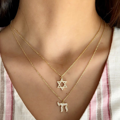 A close-up of a person's neck and chest wearing two layered gold necklaces: a Star of David pendant on the upper chain and a Hebrew letter pendant on the lower chain, both adorned with small clear stones.