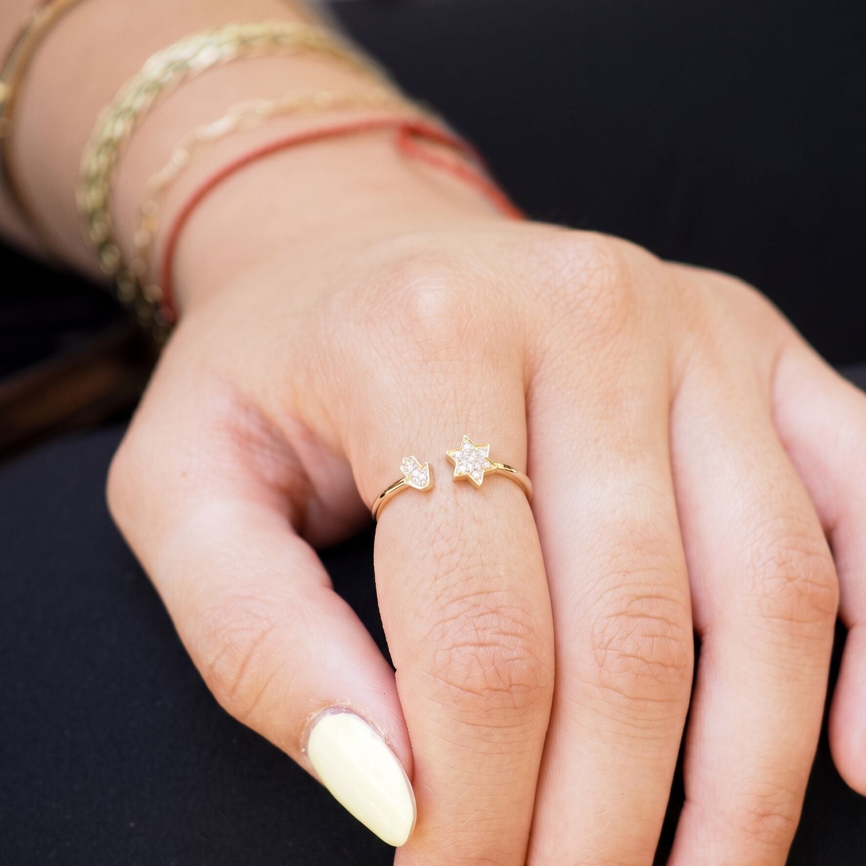 A close-up of a person's hand wearing a delicate gold ring with a small star and heart design, paired with pale yellow nail polish and gold bracelets on the wrist.