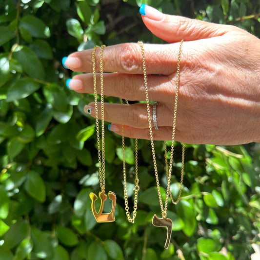 Hand holding three gold necklaces with unique pendants against a green leafy background