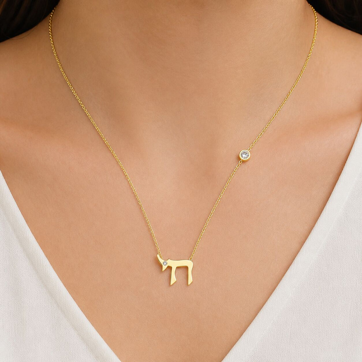 A delicate gold necklace worn by a model, featuring a small gold Hebrew letter pendant and a small round clear stone accent on the chain.