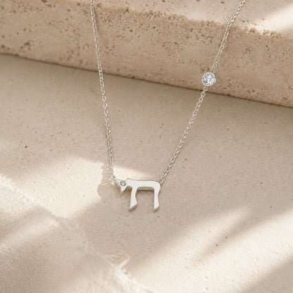 A delicate silver necklace featuring a Hebrew letter pendant and a small round bezel-set clear stone, displayed on a textured beige stone surface with soft shadows.