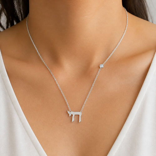 A close-up of a delicate silver chain necklace worn by a model. The necklace features a small silver chai pendant and a small round clear stone accent on the right side of the chain, set against a white V-neck top.