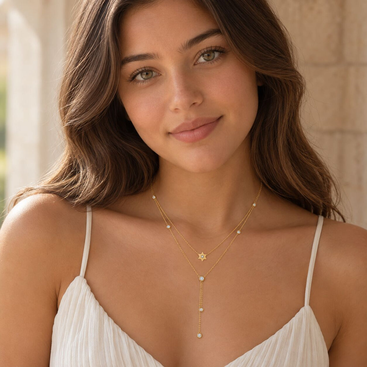 A woman wearing a delicate gold double-layer necklace with a small jewish star charm and a long drop chain accented with tiny white beads, paired with a white camisole top.
