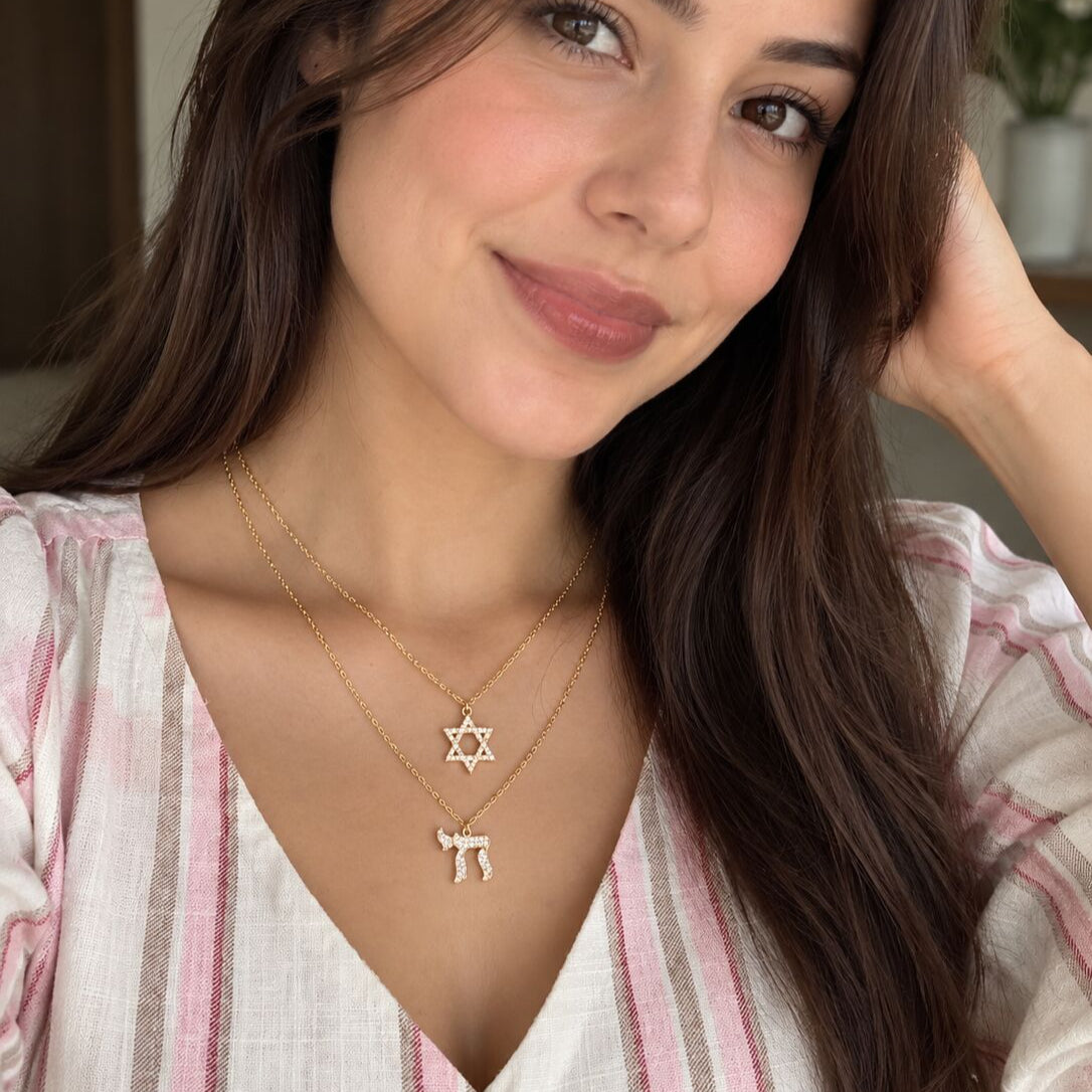 A close-up lifestyle shot of a woman wearing a double-layered gold necklace set. The top layer features a delicate chain with a Star of David pendant, and the bottom layer features a matching chain with a small donkey pendant.