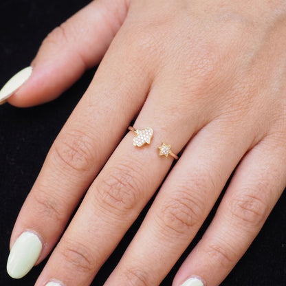 A close-up of a hand wearing two delicate gold rings against a black background. The larger ring features a textured, flower-like cluster of small clear stones, while the smaller ring is a simple gold band with a tiny clear stone.