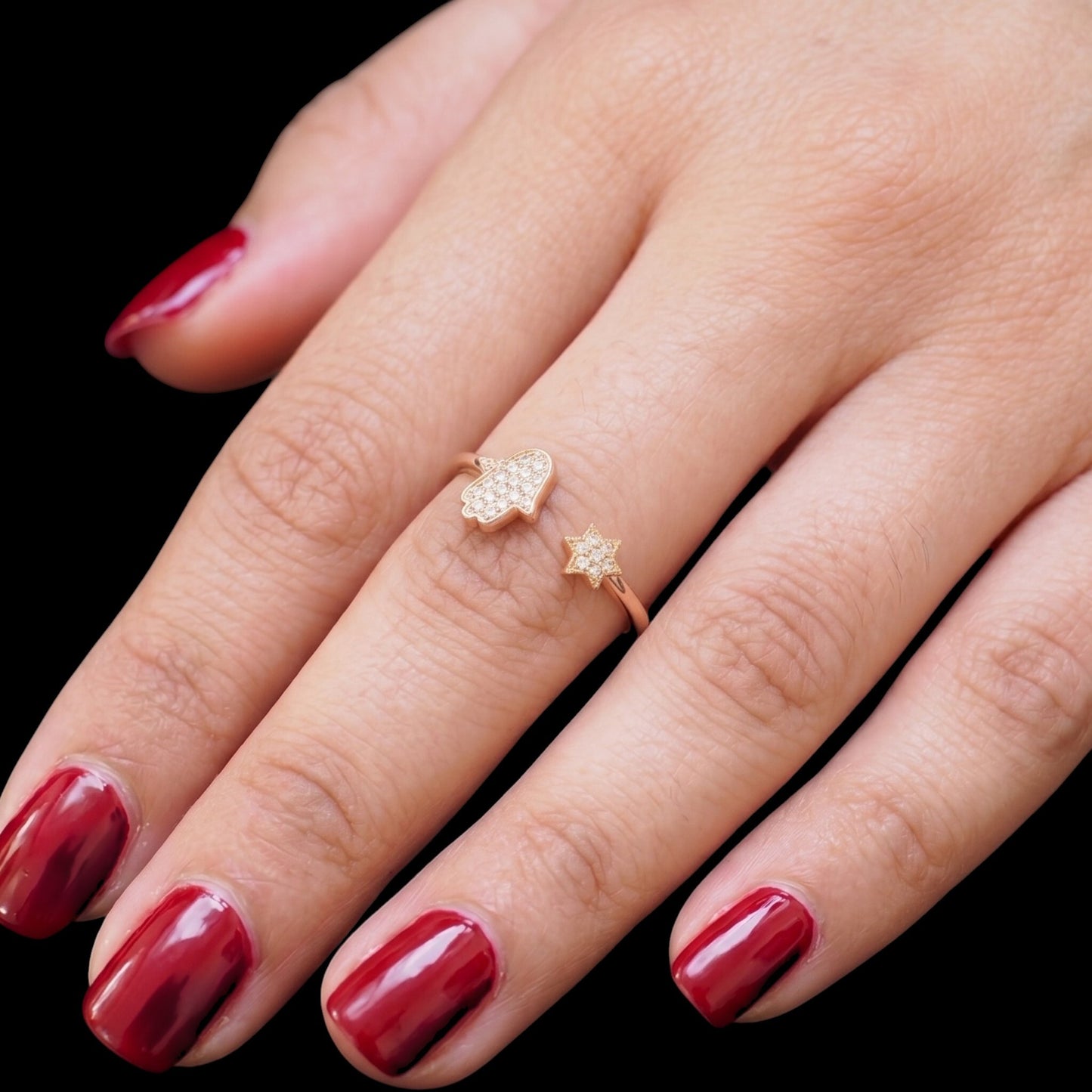 A close-up of a hand with glossy dark red nail polish wearing two delicate gold rings, one shaped like a Hamsa hand and the other like a small star, set against a solid black background.