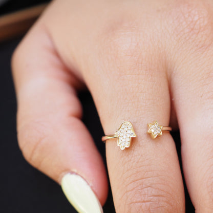 A close-up of a hand wearing two delicate gold rings: one shaped like a hamsa hand and the other like a small star, both set with tiny clear stones.