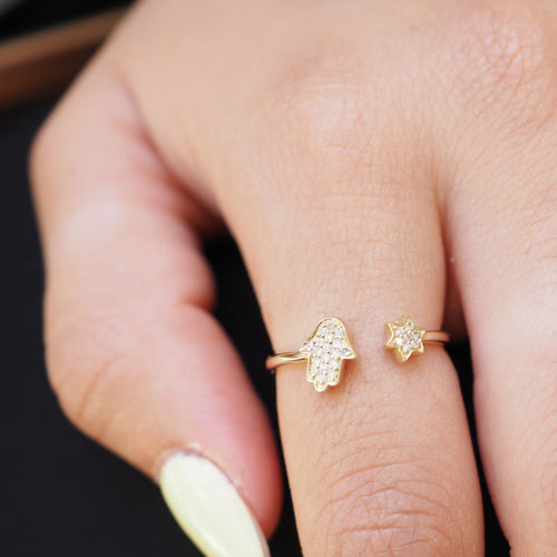 A close-up of a hand wearing two delicate gold rings: one shaped like a hamsa hand and the other like a small star, both set with tiny clear stones.