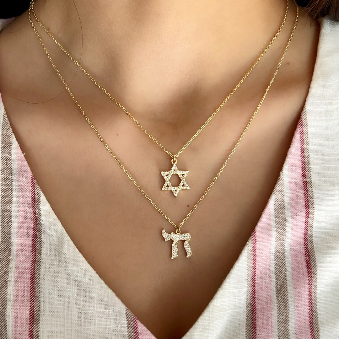 A close-up of a person wearing two delicate gold necklaces layered on the chest. The shorter necklace features a Star of David pendant, and the longer necklace features a Hebrew letter pendant (Yud).