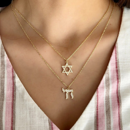 A close-up of a person wearing two delicate gold necklaces layered on the chest. The shorter necklace features a Star of David pendant, and the longer necklace features a Hebrew letter pendant (Yud).