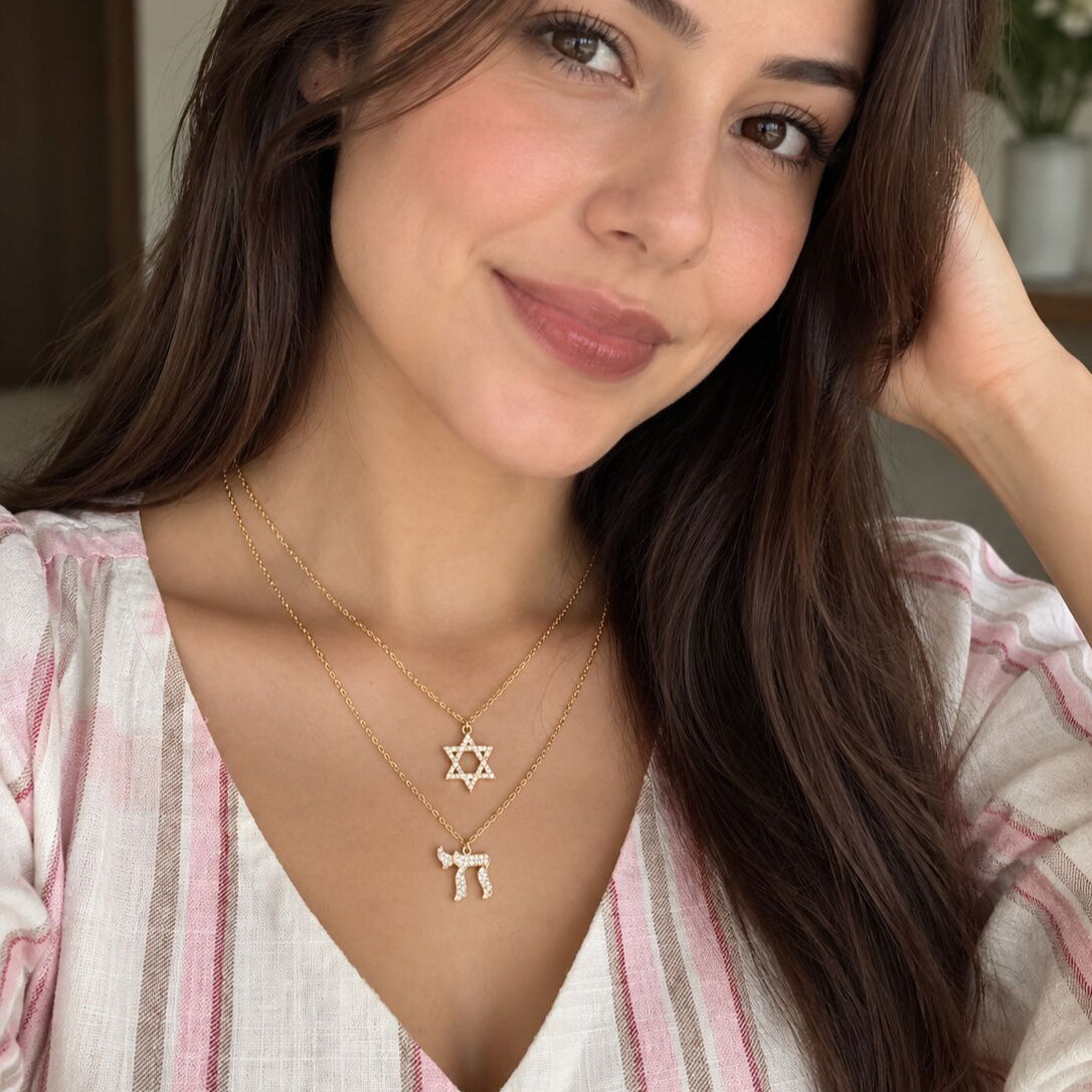 A close-up lifestyle shot of a woman wearing two delicate gold necklaces: a Star of David pendant and a Hebrew letter 'Shin' pendant, styled with a pink and white striped top.