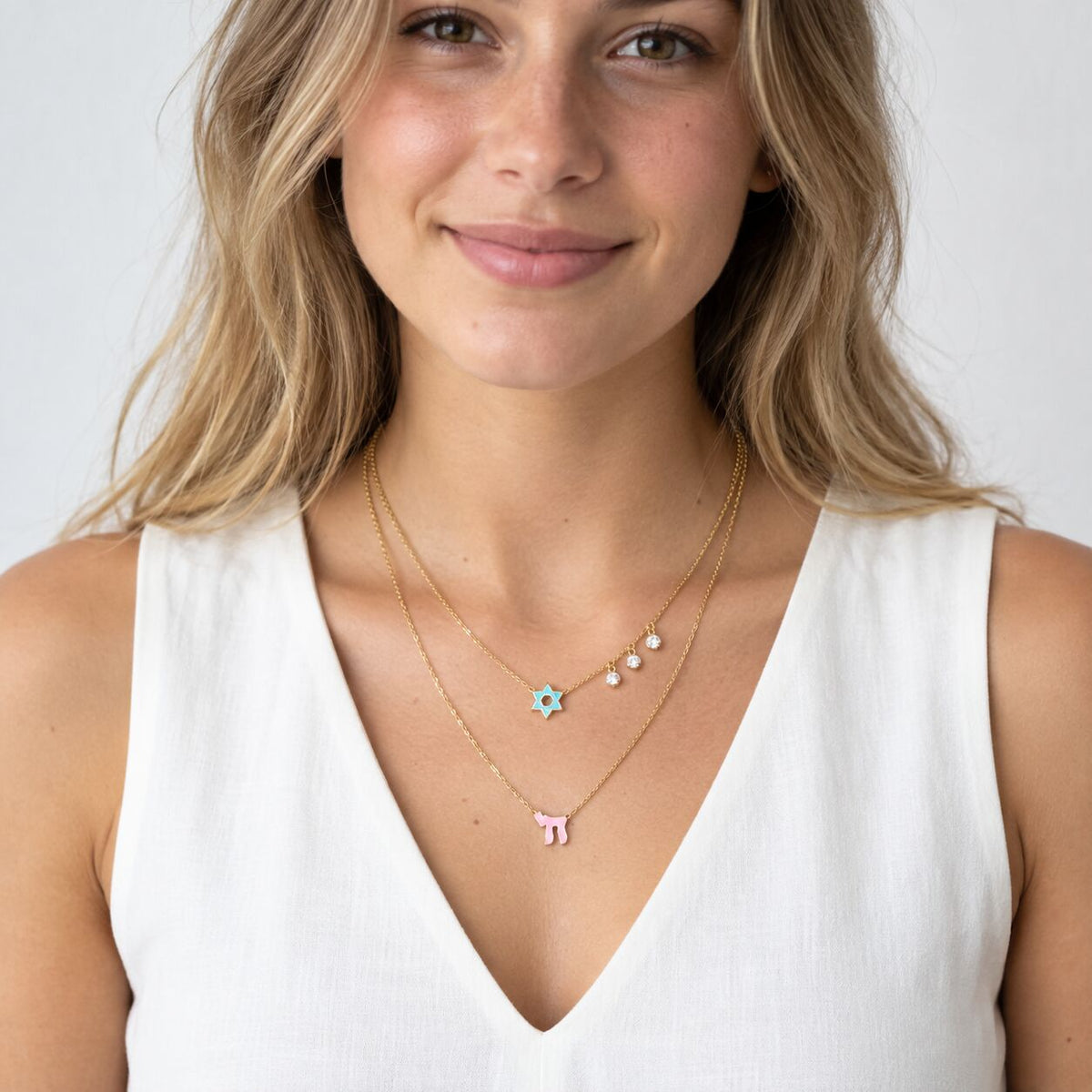 A layered necklace featuring two chains with a pink enamel Star of David and Chai symbol, accented by sparkling gems for a timeless look.